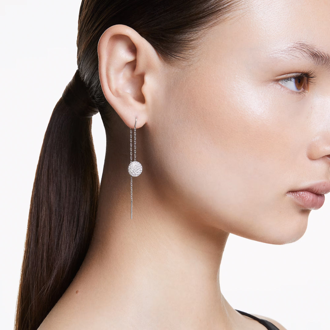 A woman with straight brown hair tied back wears a pair of Swarovski Sublima Drop Earrings. These elegant rhodium-plated earrings, adorned with crystals and featuring a spherical design alongside a short chain and slim metal stick, create a minimalist look against a plain white background.