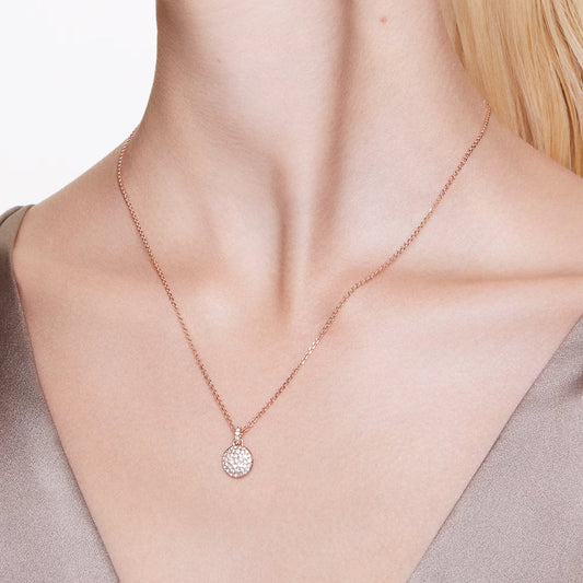A woman is wearing the Swarovski Sublima Necklace Pendant, a rose gold-tone plated necklace featuring a galaxy-inspired pendant embellished with clear round crystals. The elegant jewelry piece sits gracefully on her collarbone over a beige satin top, set against an off-white blurred background.