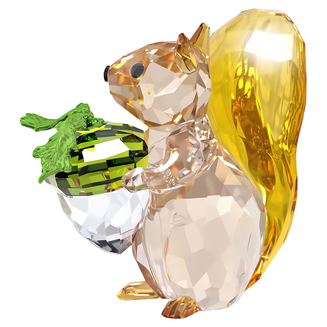 The Swarovski Idyllia Squirrel and Acorn Home Décor piece showcases a squirrel figurine holding a green acorn, with its transparent body and golden tail beautifully reflecting light.