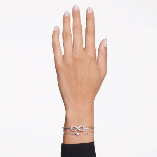 A hand with a black sleeve displays two rhodium-plated bracelets from Swarovski on the wrist. One bracelet features an infinity symbol, while the other is a simple chain embellished with Swarovski Zirconia, set against a white background.