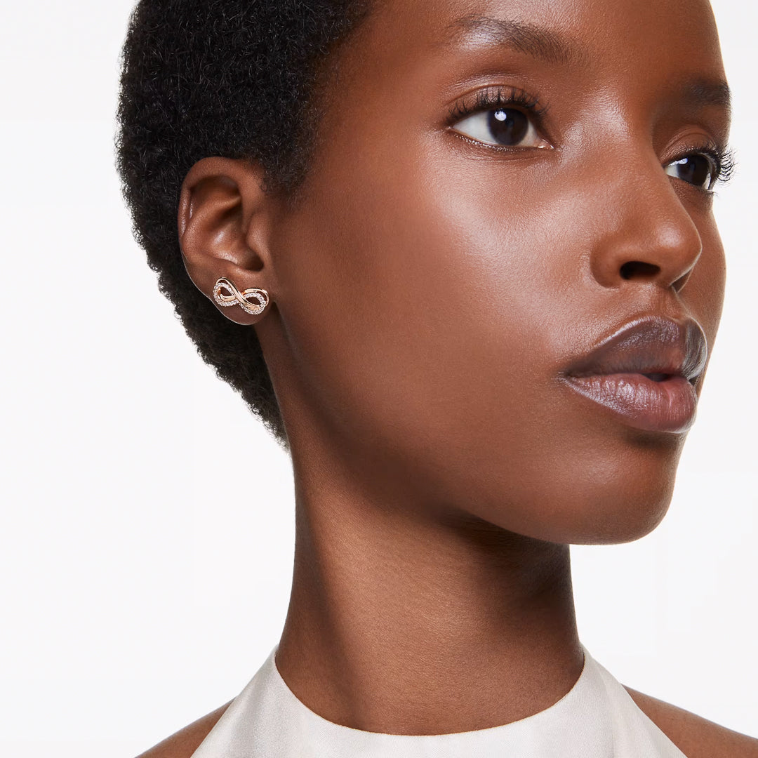 A person with short, curly hair is depicted wearing a white top along with Swarovski's Hyperbola Stud Earrings, elegantly plated in rose gold-tone. They exude a calm demeanor while gazing slightly to the left against a simple white background, embodying an essence of understated sophistication.