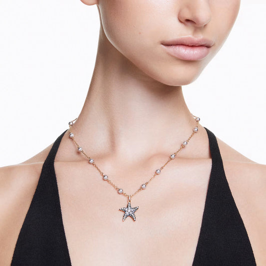 Close-up of a person wearing a black halter top and the Swarovski Idyllia Necklace Pendant adorned with crystal pearls. The gold-tone plated accessory elegantly hangs around their neck against a light background, highlighting its delicate beauty.
