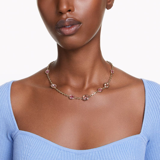 A person showcases a Swarovski Imber Necklace adorned with octagon-cut pink crystals. They are dressed in a light blue, off-the-shoulder top against a plain white background.