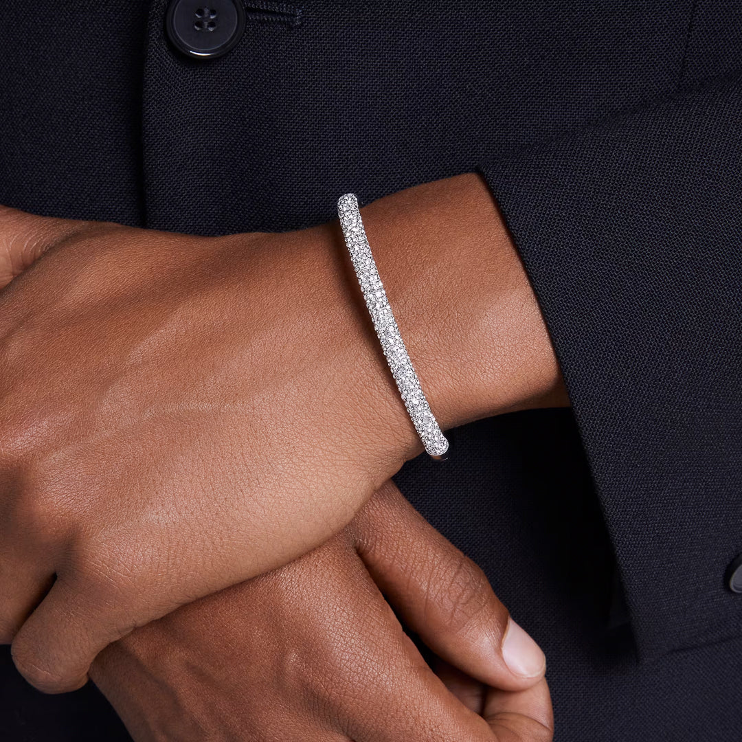 Close-up of a person wearing the elegant, silver Swarovski Sublima Bangle adorned with sparkling clear round crystals on their wrist. The rhodium-plated band shines brightly against the dark jacket as their hand gently rests on their forearm.
