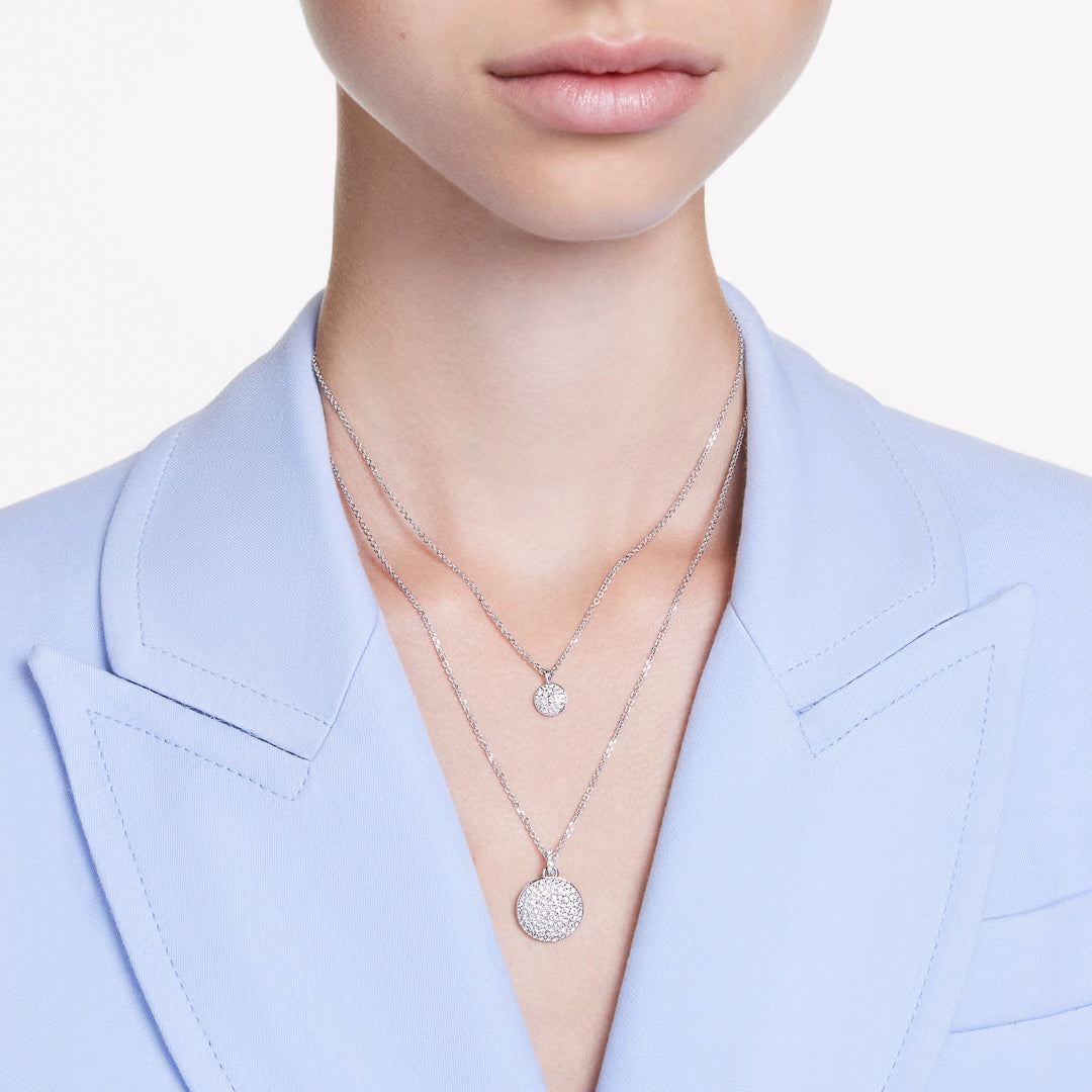 A close-up of a person wearing a light blue blazer and two silver necklaces: one features the shimmering Swarovski Sublima Layered Necklace Pendant and the other boasts a larger, textured circular design. The rhodium-plated design from Swarovski glistens against the white background.