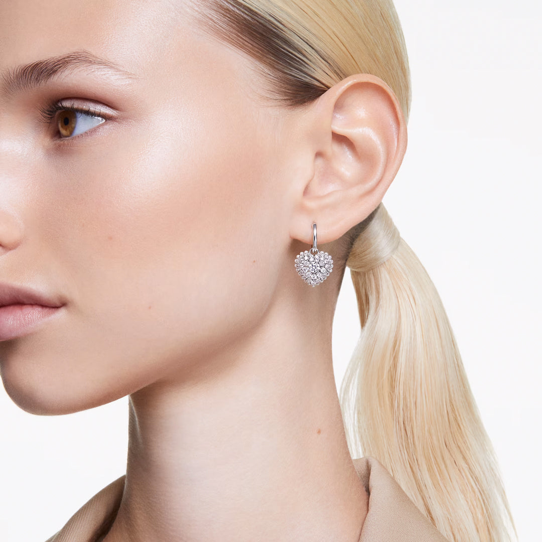 A person with blonde hair tied in a ponytail is elegantly showcasing the Swarovski Hyperbola Heart Drop Earrings, which are beautifully adorned with Swarovski Zirconia. They are wearing a beige top against a plain white background, enhancing the sparkling allure of these exquisite earrings.