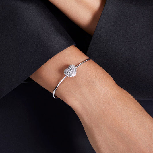 A person wearing the Swarovski Idyllia Heart Bangle, a sleek, rhodium-plated piece featuring a heart-shaped motif adorned with sparkling crystals. The bracelet rests on their wrist, partially visible against a dark fabric background.