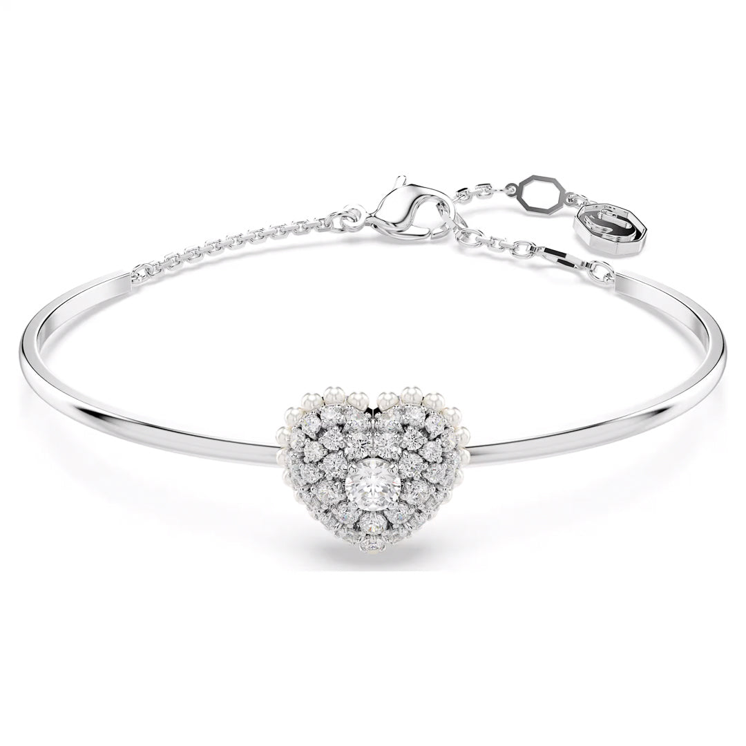 The Swarovski Idyllia Heart Bangle, crafted by Swarovski, is a delicate silver bracelet that features a heart-shaped motif embellished with multiple Swarovski Zirconia gems. It includes a thin chain with an adjustable clasp and a small tag, offering both elegance and allure in its central design.