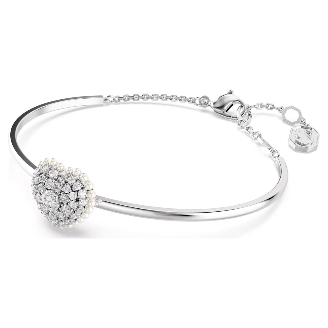 The Swarovski Idyllia Heart Bangle by Swarovski is an elegant silver bracelet with a heart-shaped motif adorned with dazzling diamonds and shimmering Swarovski Zirconia. It features an adjustable chain and a small charm at the clasp.