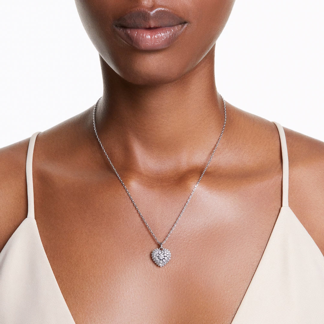 A woman in a light beige sleeveless top is adorned with the Swarovski Idyllia Heart Necklace Pendant from Swarovski, featuring a heart-shaped pendant encrusted with sparkling stones, adding an elegant touch to her outfit.