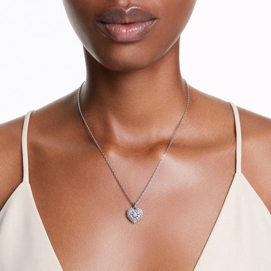 A woman in a light beige sleeveless top is adorned with the Swarovski Idyllia Heart Necklace Pendant from Swarovski, featuring a heart-shaped pendant encrusted with sparkling stones, adding an elegant touch to her outfit.
