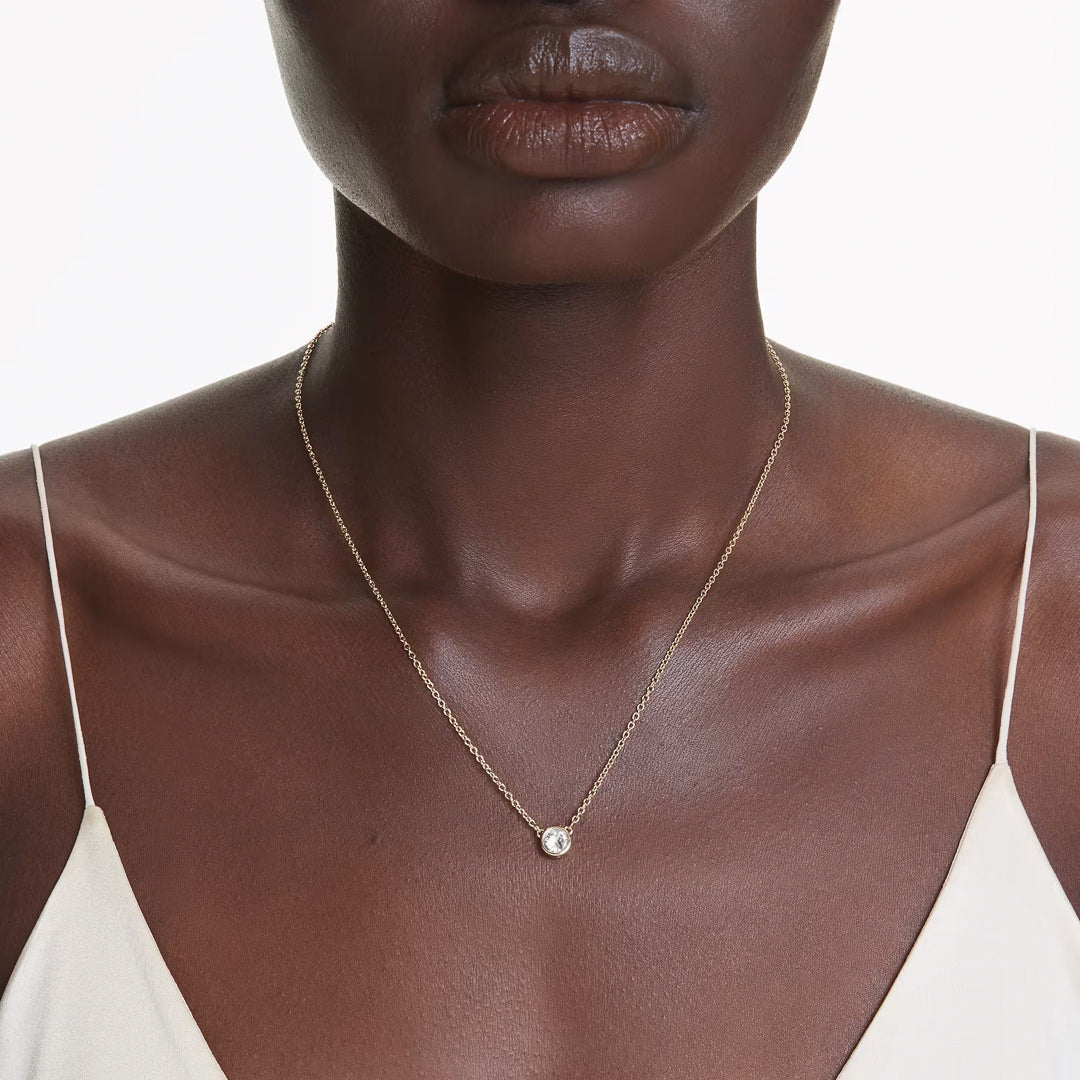 Close-up of a person wearing the Swarovski Imber Necklace Pendant, featuring gold-tone plating and set against a white background. The individual is dressed in a light-colored top with thin straps, highlighting the delicate piece embellished with sparkling Swarovski Zirconia.