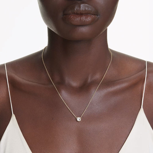 Close-up of a person wearing the Swarovski Imber Necklace Pendant, featuring gold-tone plating and set against a white background. The individual is dressed in a light-colored top with thin straps, highlighting the delicate piece embellished with sparkling Swarovski Zirconia.