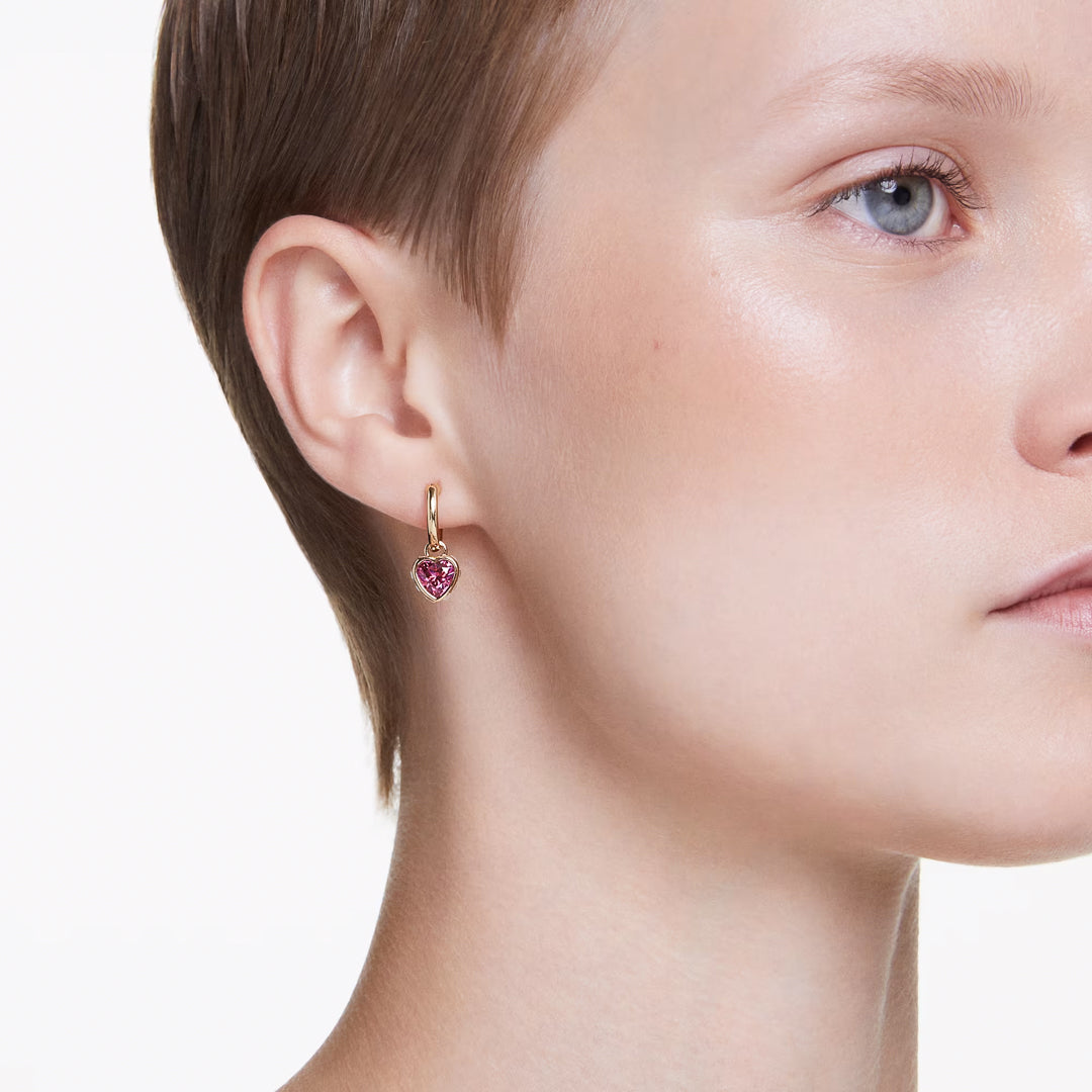 A short-haired individual is captured in profile, showcasing a luxurious gold-tone earring embellished with a heart motif featuring a pink gemstone. Their fair complexion and light blue eyes stand out, while the plain white background highlights the sophistication of this Swarovski Chroma Heart Drop Earrings piece by Swarovski.