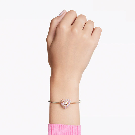 A hand clad in a pink sweater gracefully showcases the Swarovski Idyllia Heart Bangle, embellished with a dazzling heart-shaped charm inlaid with Swarovski Zirconia. The sophisticated bracelet is set against a simple white background.