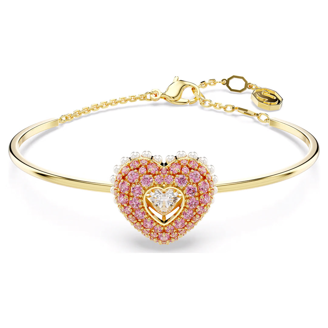 The Swarovski Idyllia Heart Bangle features a heart-shaped centerpiece adorned with pink and white gemstones. It includes a smaller gold heart accented by pink gems, layered elegantly over the design. The bracelet is embellished with Swarovski Zirconia, finished off with a delicate chain, clasp, and charm for added elegance.