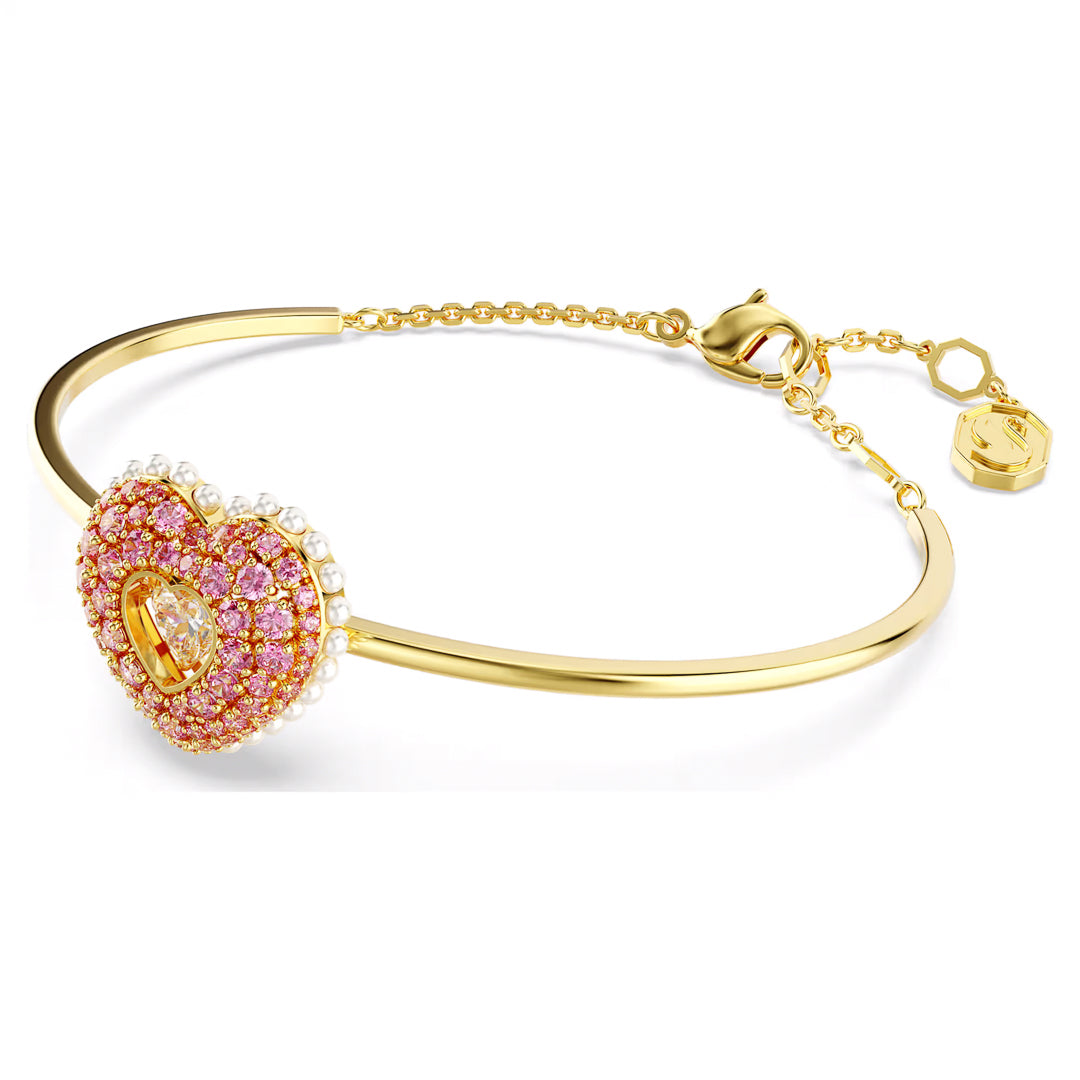 The Swarovski Idyllia Heart Bangle is a gold bracelet with a heart-shaped pendant embellished with pink and white Swarovski Zirconia stones. It features a delicate chain and an adjustable clasp, exuding elegance.