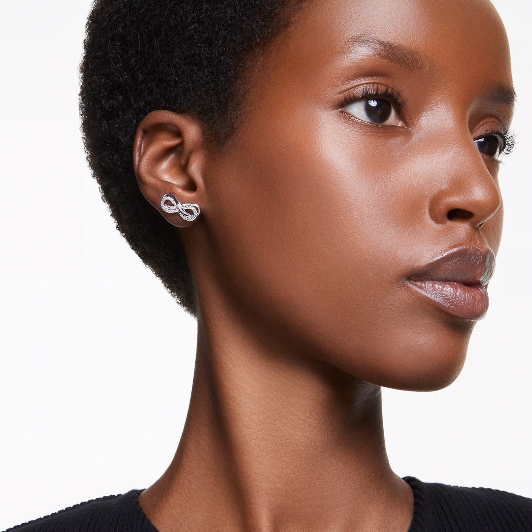 A person with short hair wears the Swarovski Hyperbola Stud Earrings, featuring rhodium plating and infinity shapes enhanced with Swarovski Zirconia. The plain white background accentuates their smooth, glowing skin and the jewelry's elegance, while they are dressed in a black top.