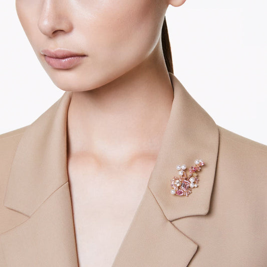 A person in a beige blazer is adorned with a Swarovski Gema Brooch, featuring pink and white Crystal Pearls by the brand Swarovski. The image focuses on the upper torso and the brooch, set against a plain white background.