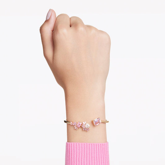 A hand adorned with the Swarovski Gema Bangle, a piece by Swarovski featuring pink gemstone embellishments reminiscent of cherry blossoms, set against a plain white background. The wrist is elegantly covered by a pink ribbed sleeve.