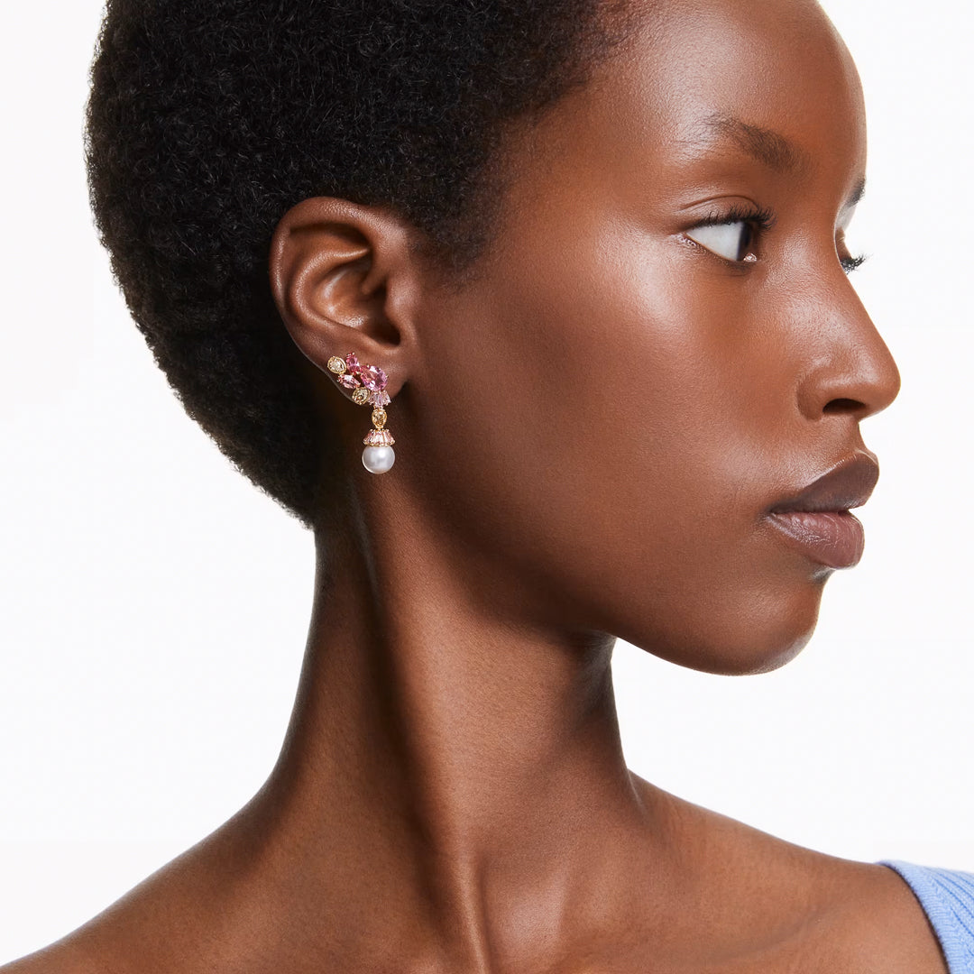 A person with short hair elegantly wears Swarovski Gema Drop Earrings adorned with pink gemstones and a dangling pearl. They are looking to the side against a plain white background, dressed in a light blue top.