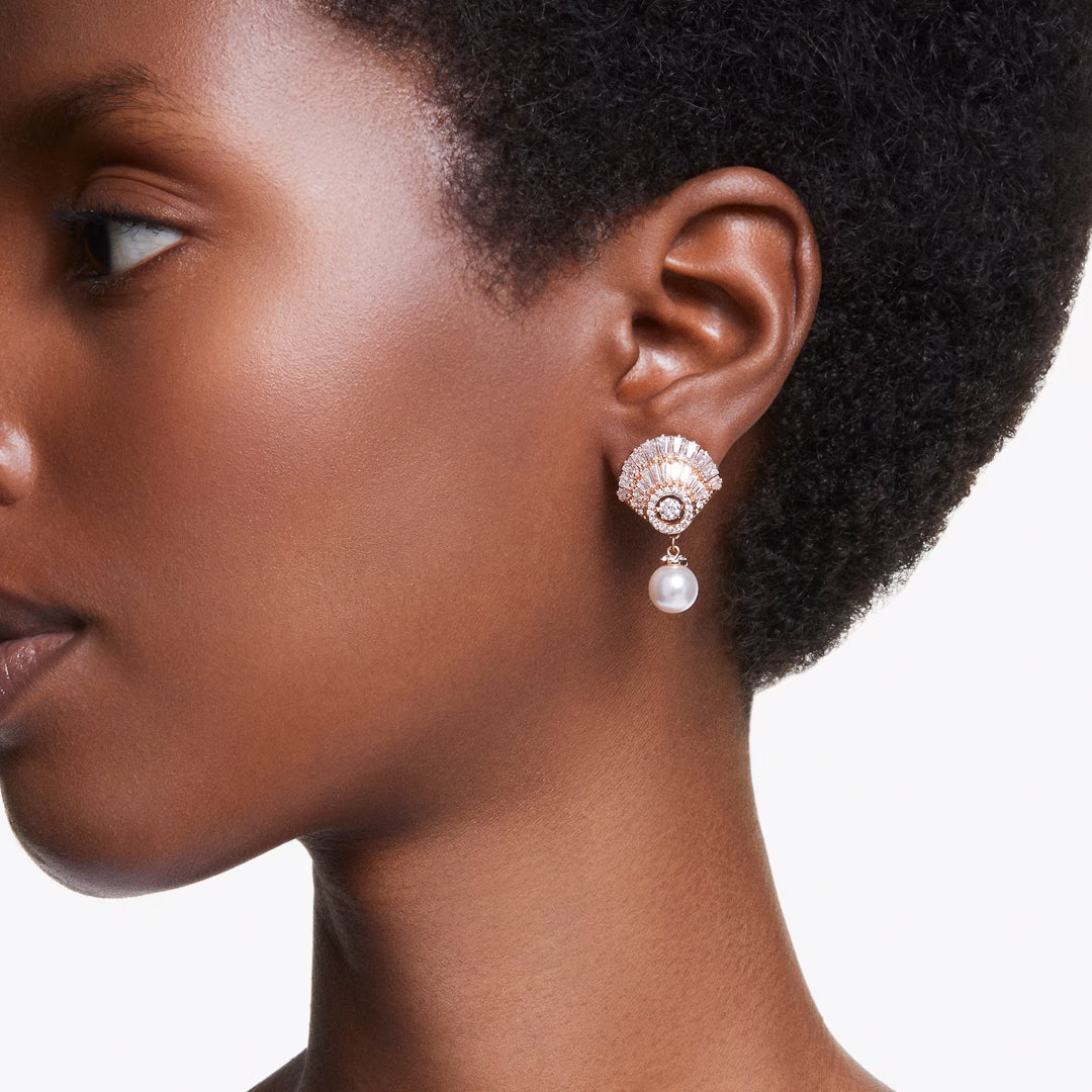 Close-up of a person wearing the Swarovski Idyllia Drop Earrings, featuring a shell-shaped design embellished with Swarovski Zirconia and a dangling pearl. The rose gold-tone plating of the jewelry shines elegantly against the person's short, curly hair and smooth skin, creating a serene backdrop.