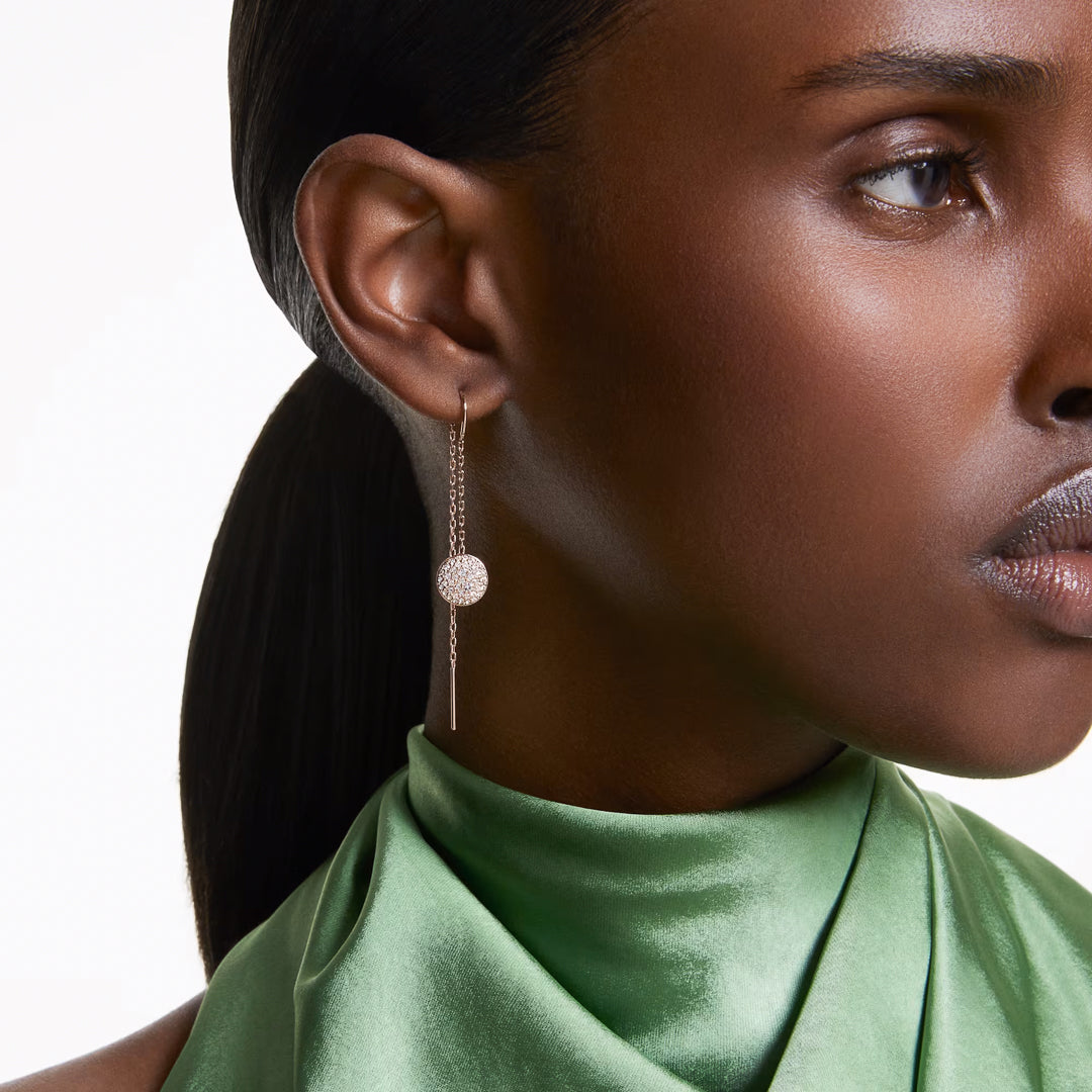 A person with a sleek ponytail wears a dangling Swarovski Sublima Drop Earring and a green satin top. The rose gold-tone earring, from Swarovski, showcases a delicate chain adorned with clear round crystals. The image captures a side profile focused on the exquisite detail of the earring.