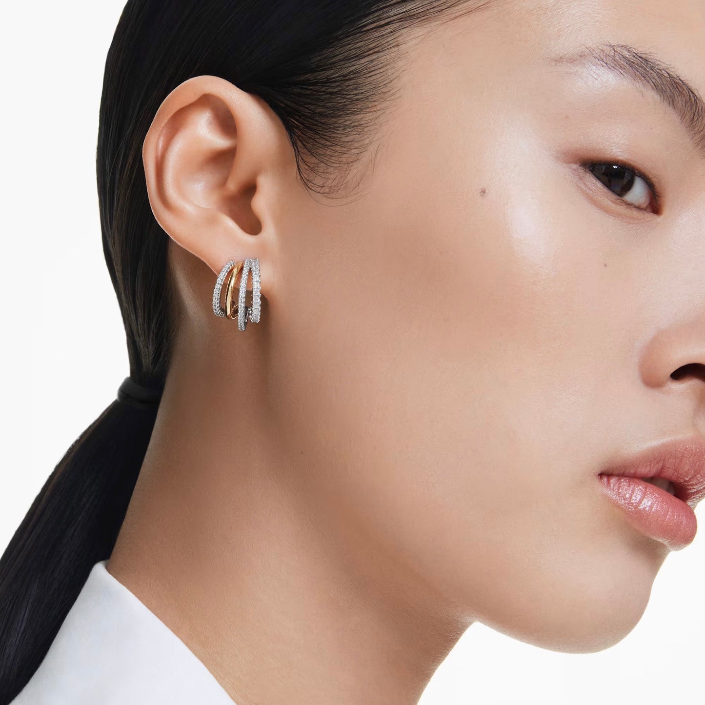 The image features a close-up of a person with sleek black hair wearing the Swarovski Hyperbola Hoop Earrings. Adorned with Swarovski Zirconia, these exquisite triple hoop diamond earrings are beautifully highlighted against the white background, enhancing both the person's flawless skin and the elegance of these stunning accessories.