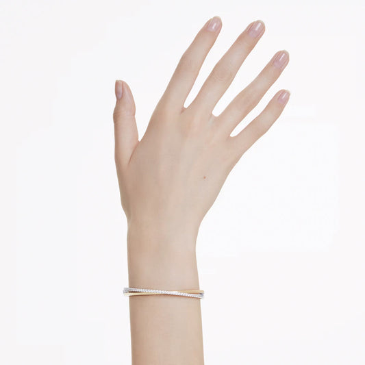 A hand with neatly manicured nails is adorned with the Swarovski Hyperbola Bangle by Swarovski. This thin, elegant bracelet features two slender, intertwined bands—one gold and one silver—and stands out beautifully against a plain white background.