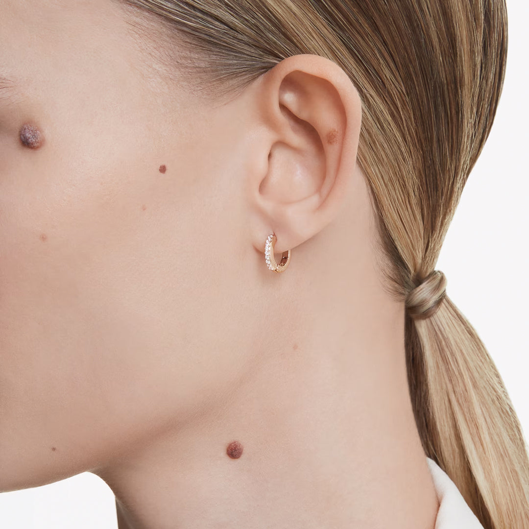 Close-up of a person with blond hair tied back, showcasing an exquisite pair of Swarovski Matrix Hoop Earrings in rose gold-tone, adorned with Swarovski Zirconia. Distinct moles appear on the cheek and neck. The person is dressed in a white collared shirt, highlighting the ear and earring.