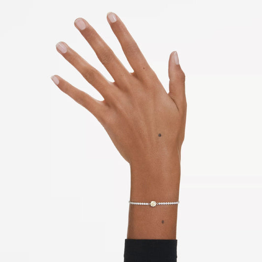 A hand with neatly manicured nails appears against a light background, its wrist elegantly adorned with the Swarovski Matrix Tennis Bracelet. The delicate bracelet from Swarovski features small round beads and a striking central golden circle, glimmering subtly while the person wears a black sleeve.