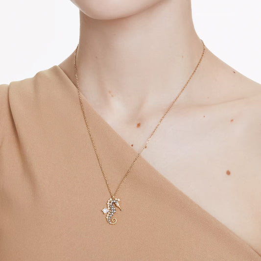 A woman wearing a tan, one-shoulder top and the Swarovski Idyllia Necklace Pendant by Swarovski, featuring an oceanic motif with a seahorse pendant adorned with sparkling Swarovski Zirconia. The background is plain white.