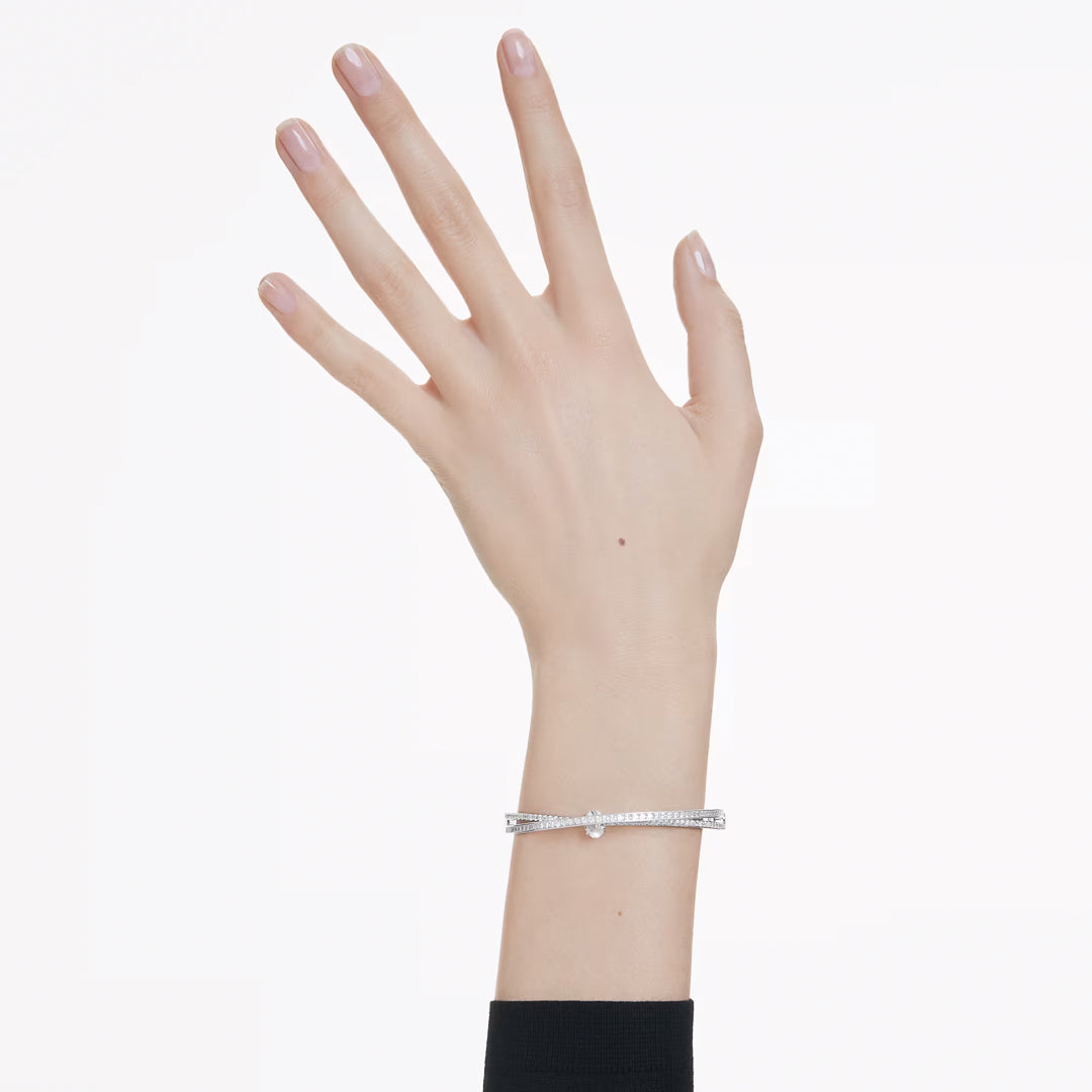 A hand with a light skin tone showcases the Swarovski Hyperbola Bangle, a rhodium-plated bracelet shaped like a miniature nail. The manicured nails are adorned with light pink polish, all set against a plain white background.