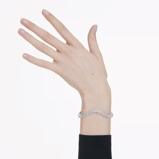 Against a white background, a hand with neatly manicured nails displays the exquisite Swarovski Sublima Bangle, embellished with small sparkling stones. The person is dressed in a black long-sleeve top, which enhances the sophisticated ruthenium-plated finish of the bracelet.