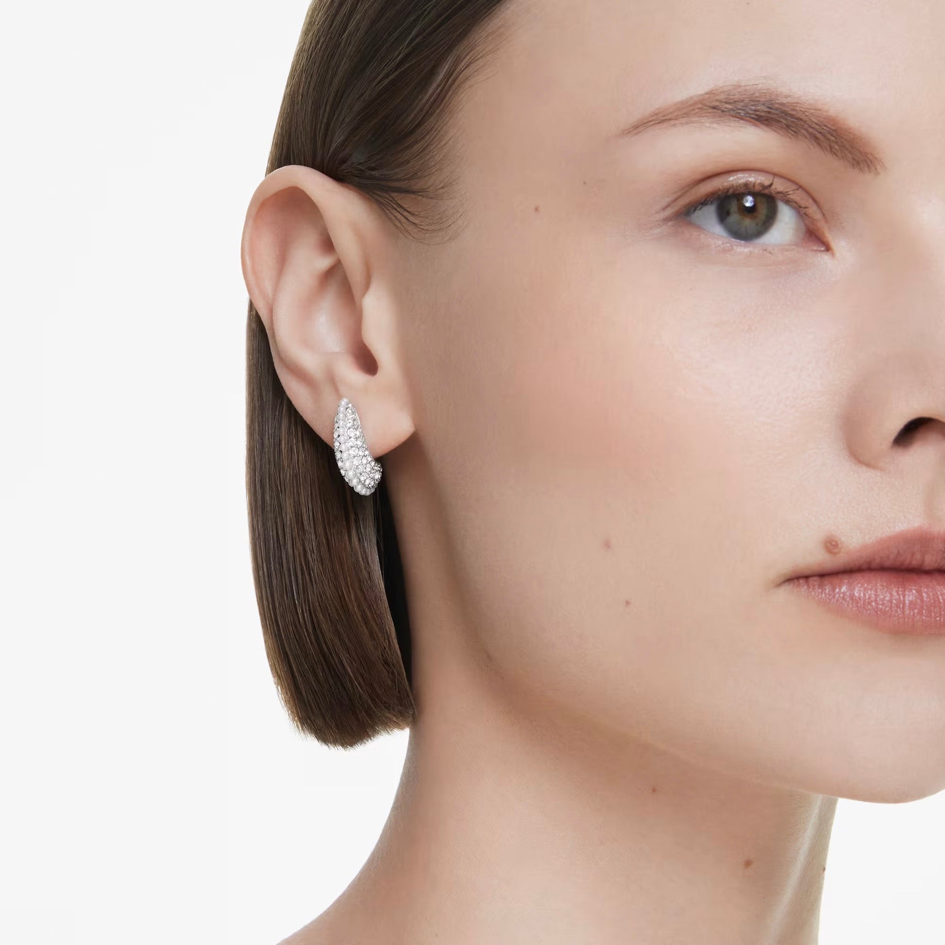 A close-up depicts a person with short brown hair wearing the Swarovski Sublima Drop Earrings, featuring a sparkling texture adorned with pavé clear crystals. Their right side is visible, highlighting their clear skin and subtle makeup against a plain white background.