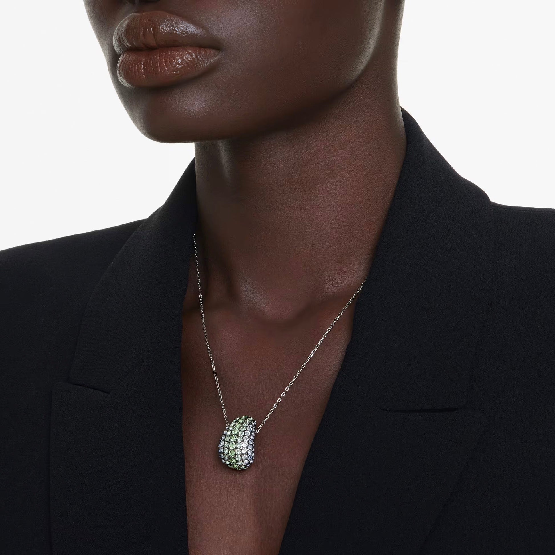 A person wearing a black blazer showcases the Swarovski Sublima Necklace Pendant, adorned with crystals in silver and marine-inspired shades. The close-up captures the elegance and intricate details of the jewelry, while the plain white background emphasizes the necklace's beauty.