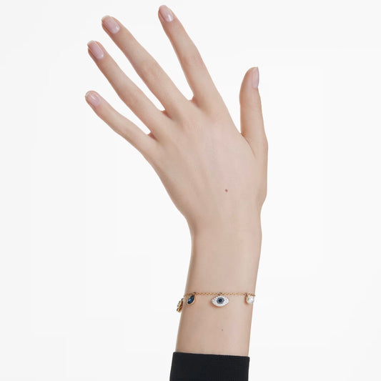 A hand with a black sleeve is displayed, showcasing the Swarovski Symbolica Charm Bracelet, a gold-tone piece featuring an eye pendant and several small charms that capture the essence of symbolic jewelry. The background is plain white.