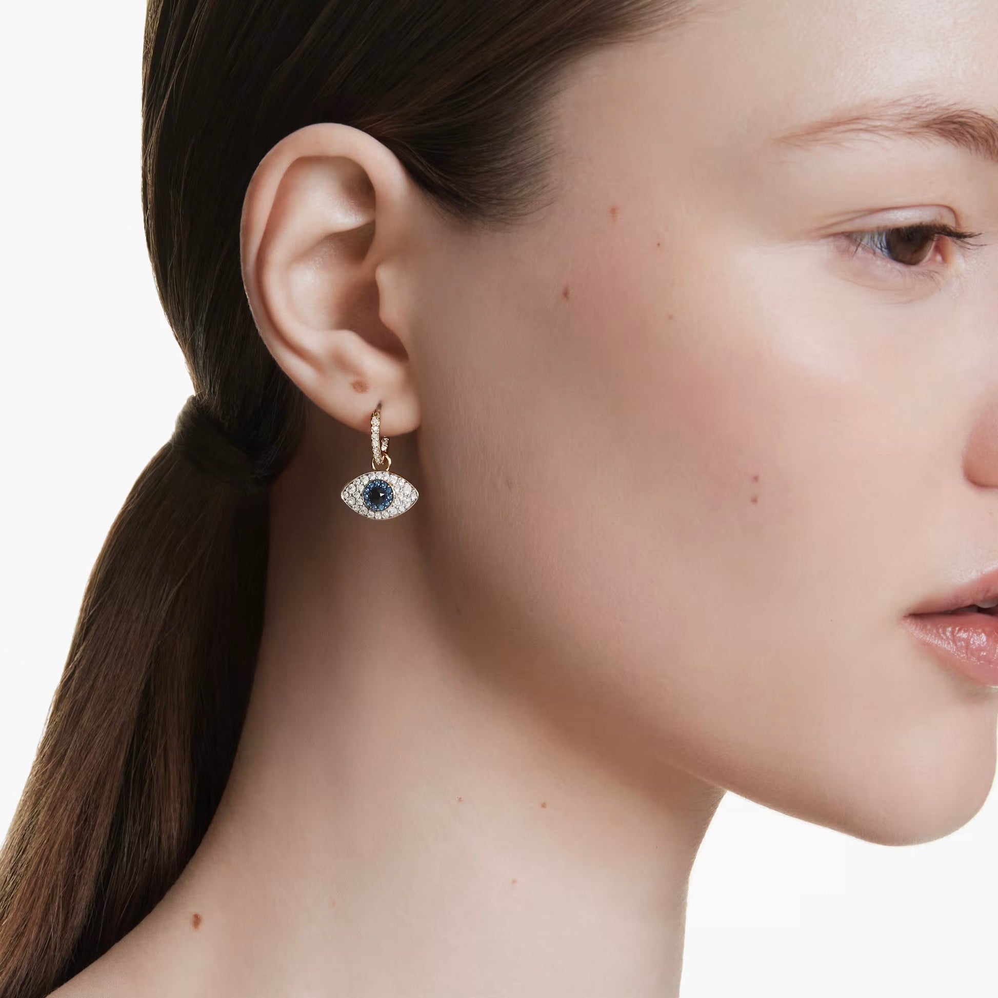 A person with long, dark hair tied back adorns themselves with the captivating Swarovski Symbolica Evil Eye Drop Earrings. This piece from Swarovski is designed as an evil eye motif, featuring a dark central stone encircled by smaller clear stones, creating a mystical glamour against the plain white backdrop.