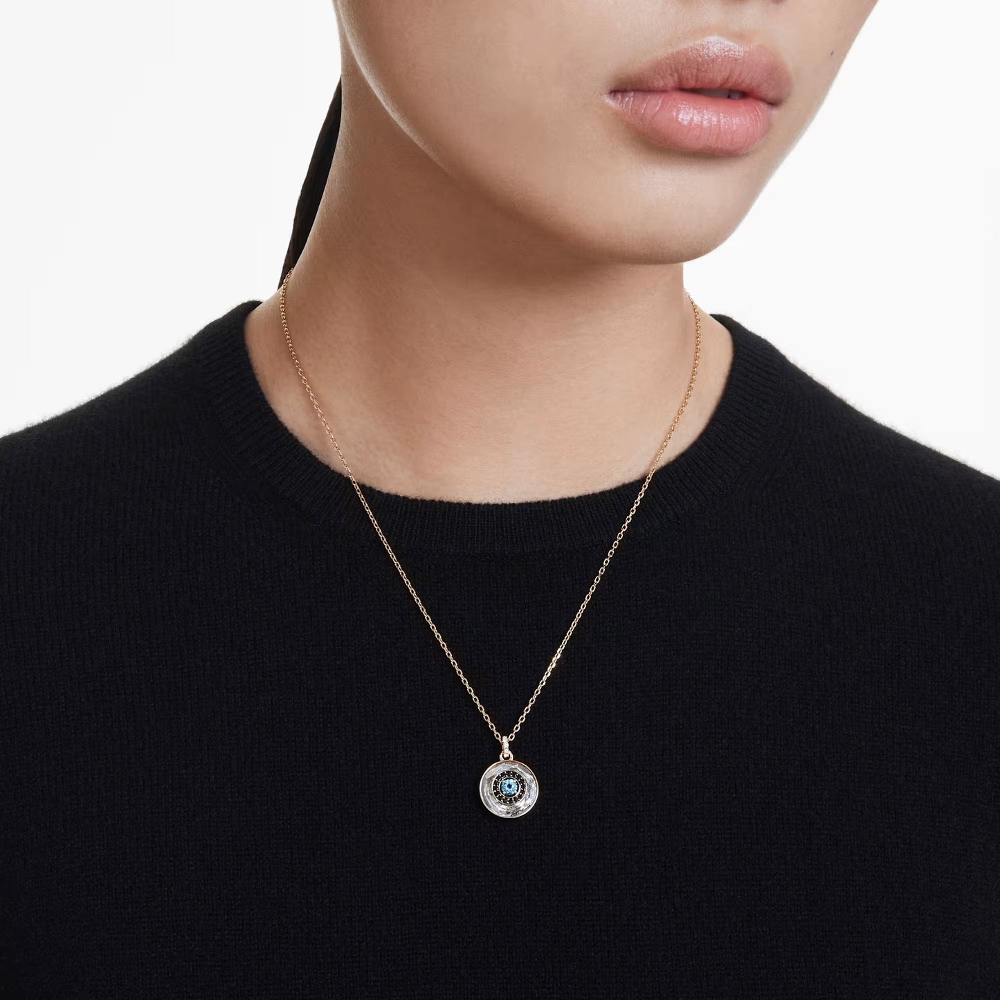 A close-up of a person wearing a black top and showcasing gold-tone jewelry, featuring the Swarovski Symbolica Evil Eye Necklace Pendant. The circular charm boasts an intricate blue and silver design.