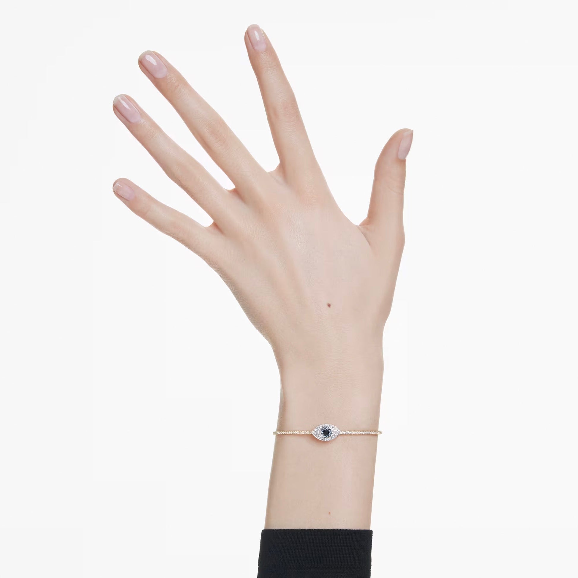 A hand adorned with a Swarovski Symbolica Evil Eye Bangle, which features an evil eye charm and a gold-tone plated chain, appears against a plain background. The person showcases neatly manicured nails peeking from a black sleeve.