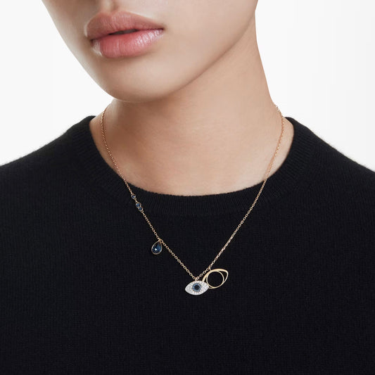 A person wearing a black sweater showcases the Swarovski Symbolica Evil Eye Necklace Pendant, featuring an eye-shaped design adorned with crystals. The necklace is completed with a small blue teardrop charm and a circular gold loop, embodying an elaborate evil eye motif by Swarovski.