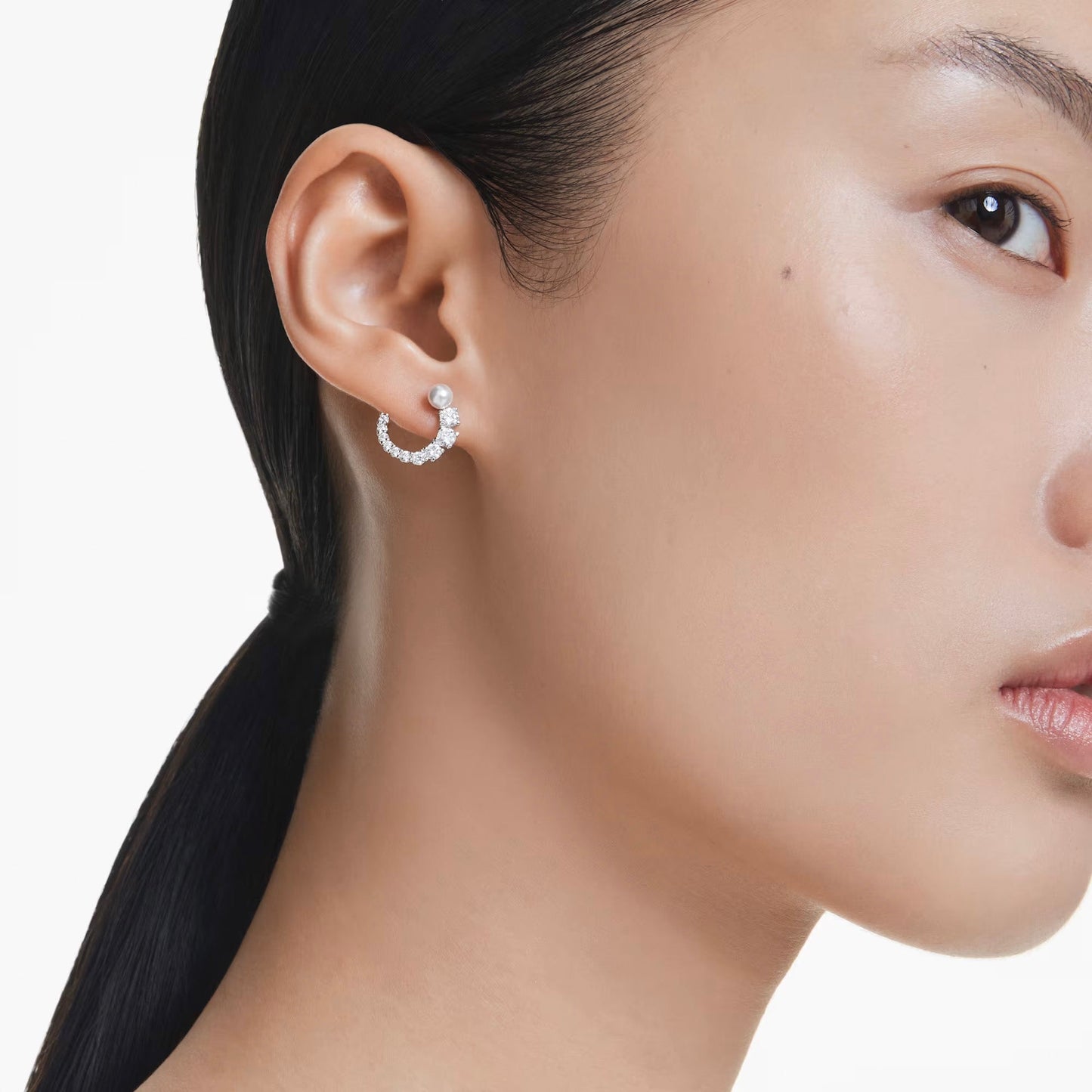 Close-up of a person with sleek black hair tied back, showcasing a shimmering pair of Swarovski Matrix Hoop Earrings, plated in rhodium and set with small Swarovski Zirconia, accompanied by a crystal pearl stud above it, against a plain white background.