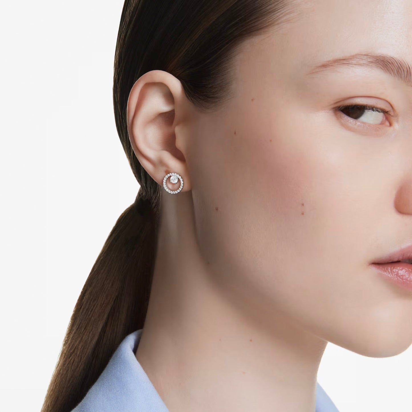 Close-up of a woman with fair skin and brown hair tied back, wearing the Swarovski Constella Stud Earrings. She is dressed in a light blue shirt, set against a plain white background.