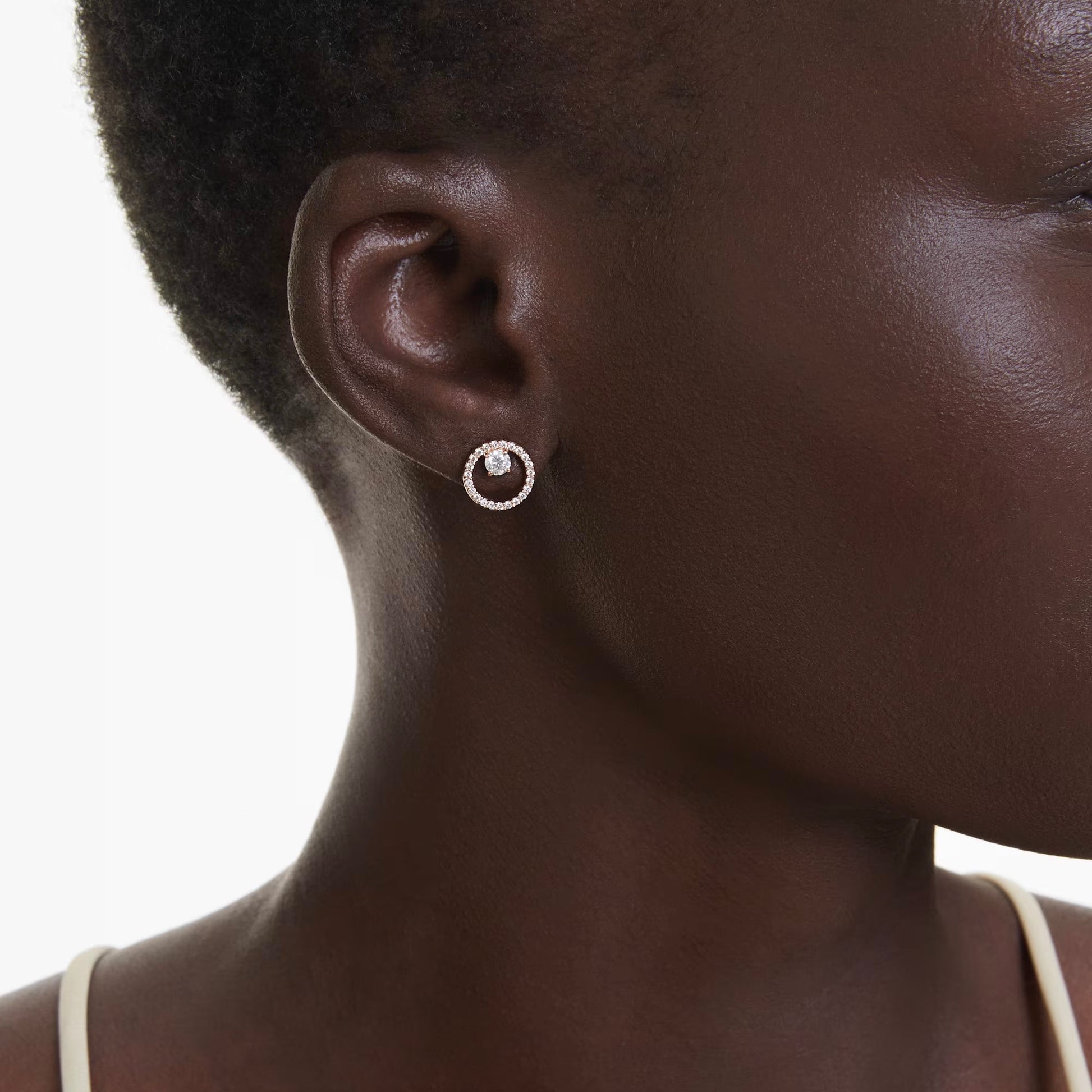 A close-up of a person's right ear showcases the Swarovski Constella Stud Earrings by Swarovski. These round stud earrings feature a central Swarovski Zirconia surrounded by an intricate design and are elegantly plated in rose gold-tone, beautifully contrasting with the white background and enhancing the person’s short hair.