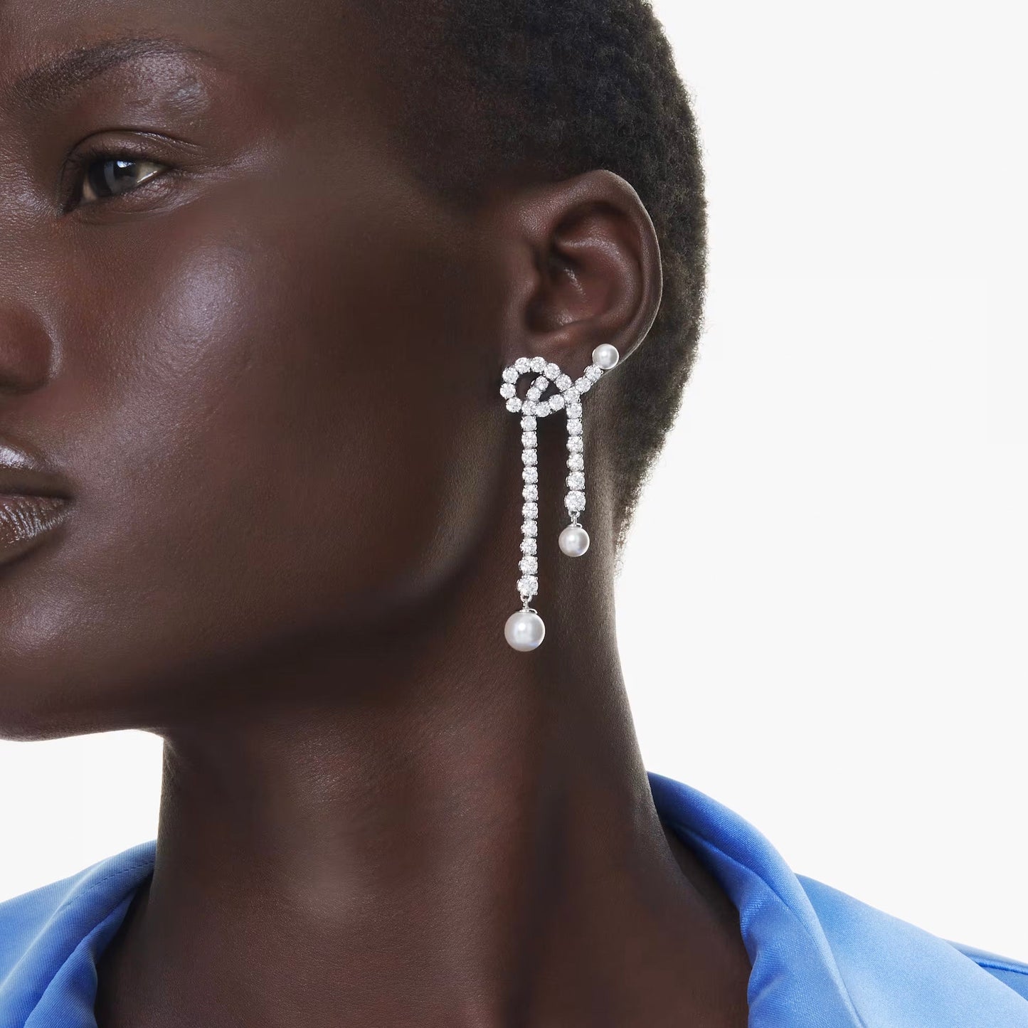 A person adorned with Swarovski Matrix Drop Earrings, featuring large and intricate designs made of Swarovski Crystal Pearls that elegantly wrap around the ear and cascade down in strands. They are dressed in a silky blue outfit against a plain white background, exuding elegance and grace.