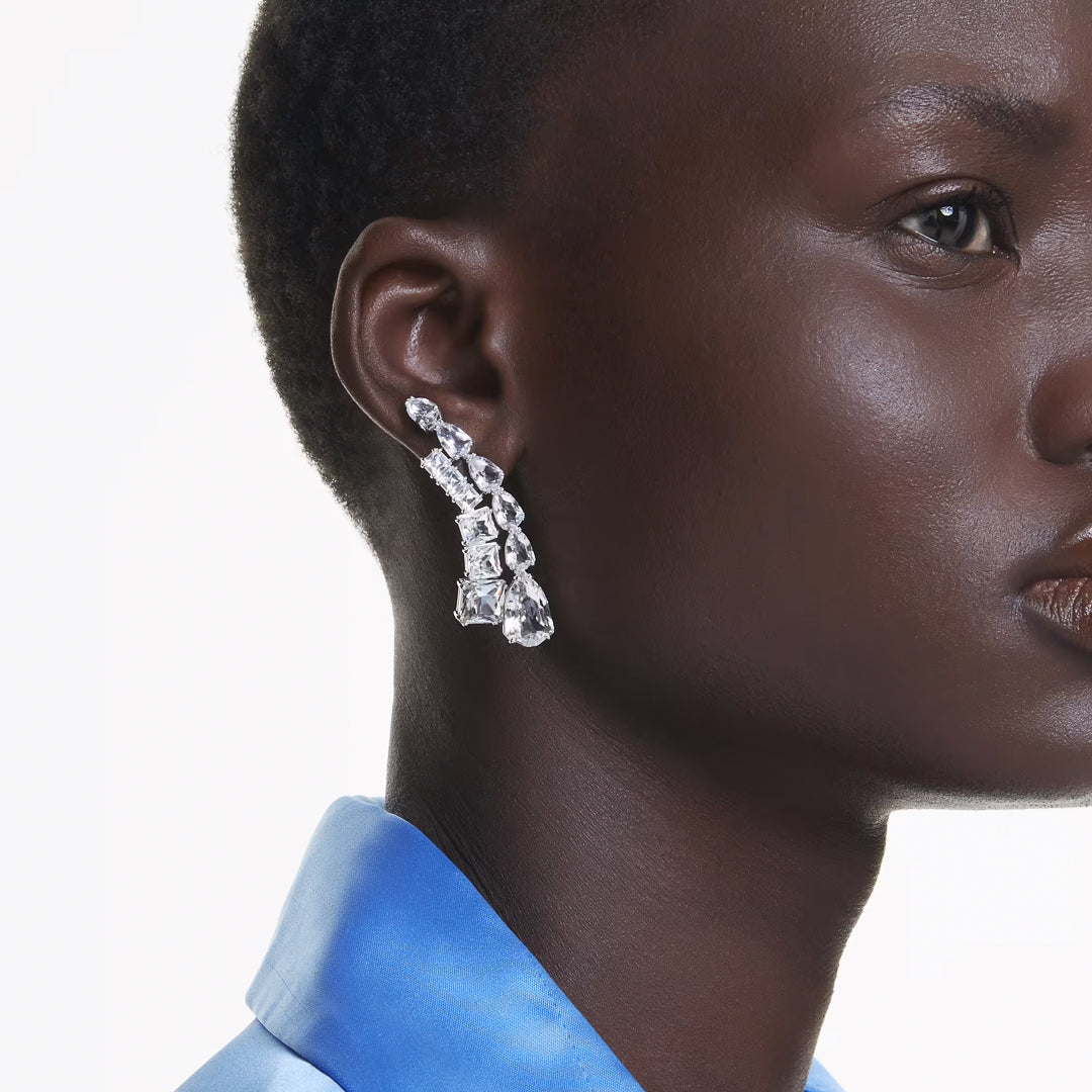 A person wearing Swarovski Matrix Drop Earrings, adorned with geometric shapes and shimmering crystals, is shown in profile. They have a smooth complexion and are dressed in a bright blue, silky collared blouse against a plain white background.