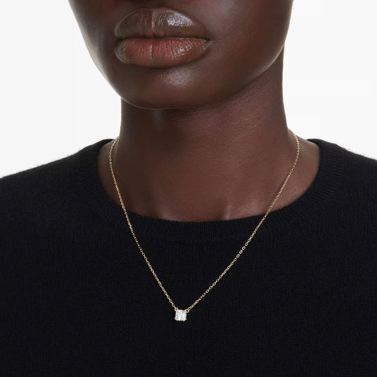 A close-up shot features an individual wearing a Swarovski Stilla Attract Necklace Pendant, showcasing its elegant gold-tone plating and small, rectangular Swarovski Zirconia charm against a black top. The person's lips and chin are visible, with a plain white background completing the scene.