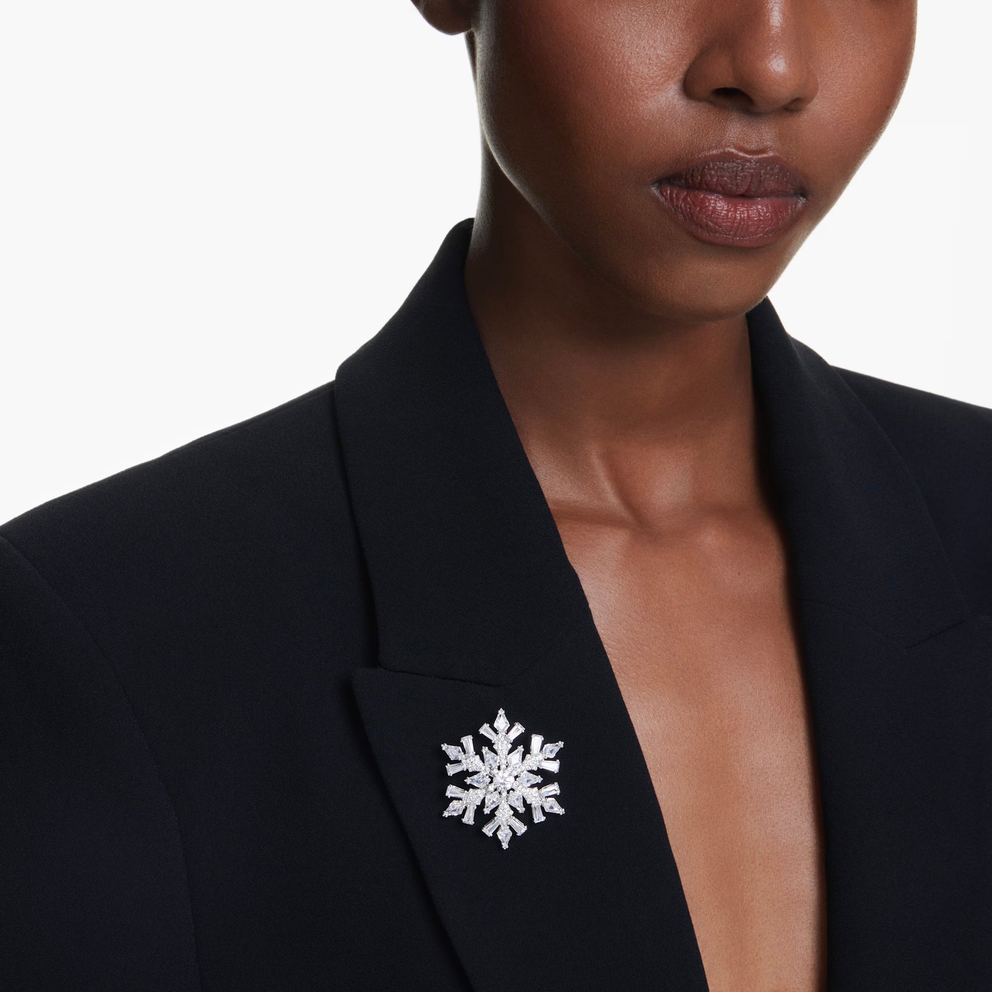 A person wearing a black blazer features the Swarovski Idyllia Necklace Pendant and Brooch on the lapel, intricately designed like a snowflake. Embellished with Swarovski Zirconia, it shines brilliantly against the white background, highlighting its elegance and creating a refined contrast with the dark fabric.
