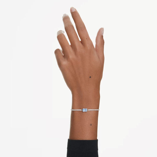 A hand adorned with the exquisite Swarovski Matrix Tennis Bracelet, showcasing a captivating blue Swarovski Zirconia centerpiece. This slender, sophisticated piece is rhodium plated for lasting durability and accented with delicate beads along its length. The hand rests gracefully against a neutral background.