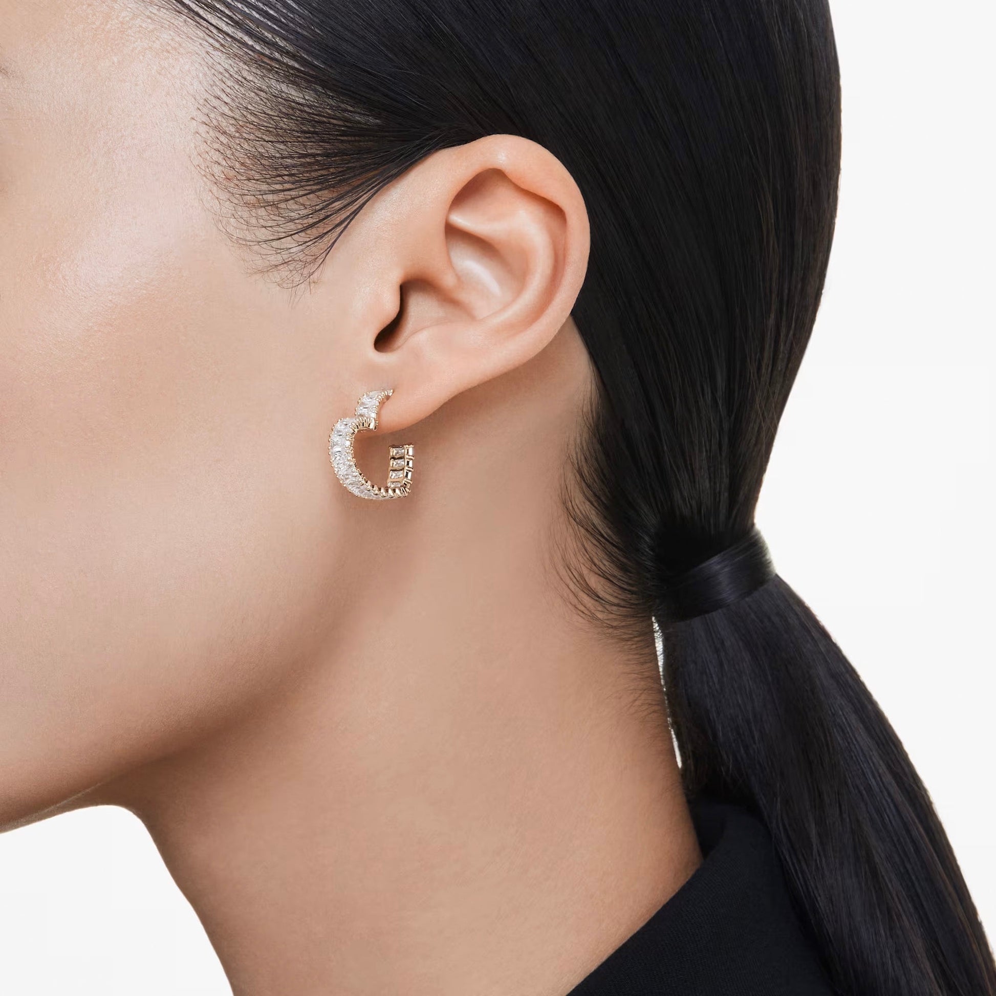 A close-up of a person wearing the sparkling Swarovski Matrix Heart Hoop Earrings, which are adorned with small stones and feature gold-tone plating. The person has straight, black hair pulled back into a low ponytail and is wearing a black top.